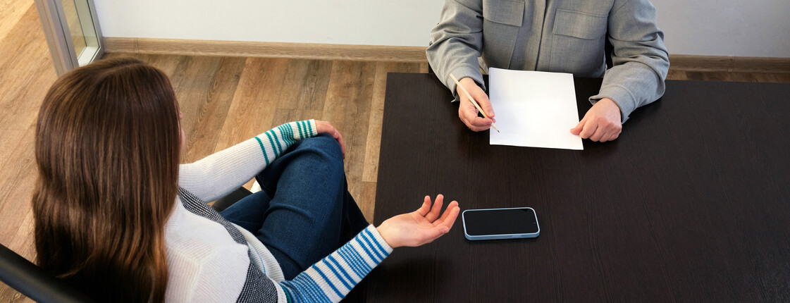Beratung für Sex-Dienstleister*innen Eine junge Frau sitzt an einem Besprechungstisch einer Frau mittleren Alters gegenüber. Auf dem Tisch liegen Schreibunterlagen.